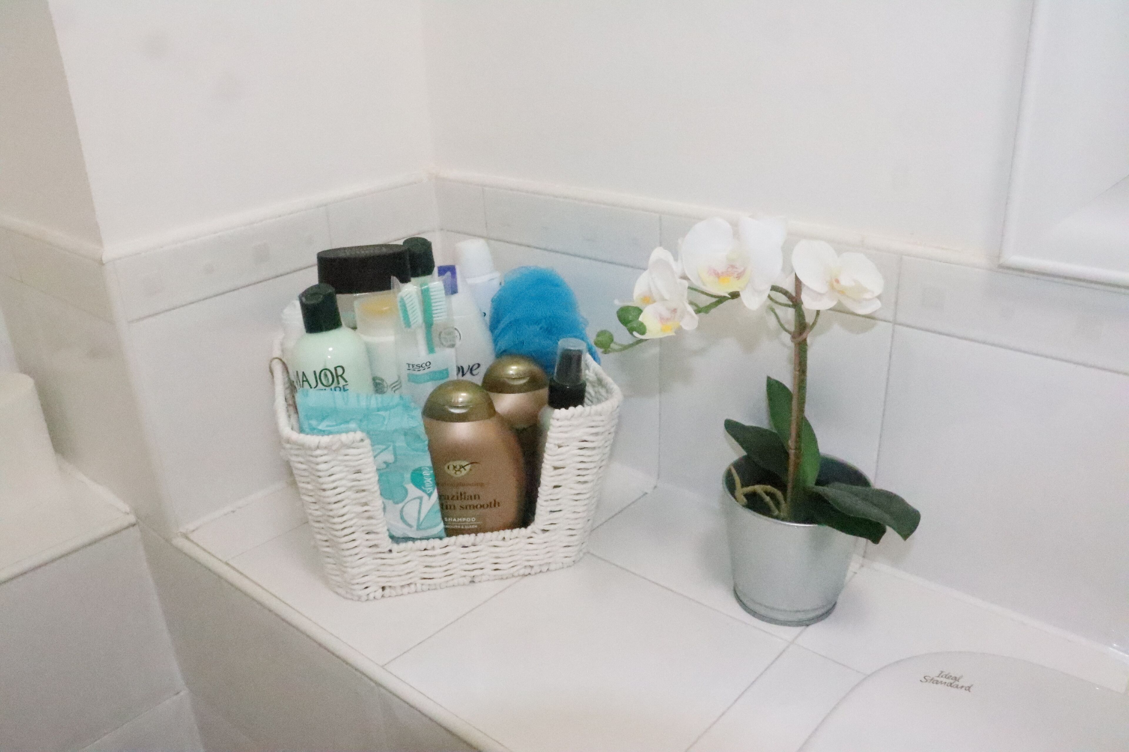 A bathroom with a white tiled wall, a white wicker basket containing toiletries, a potted plant with white flowers, and a white sink.