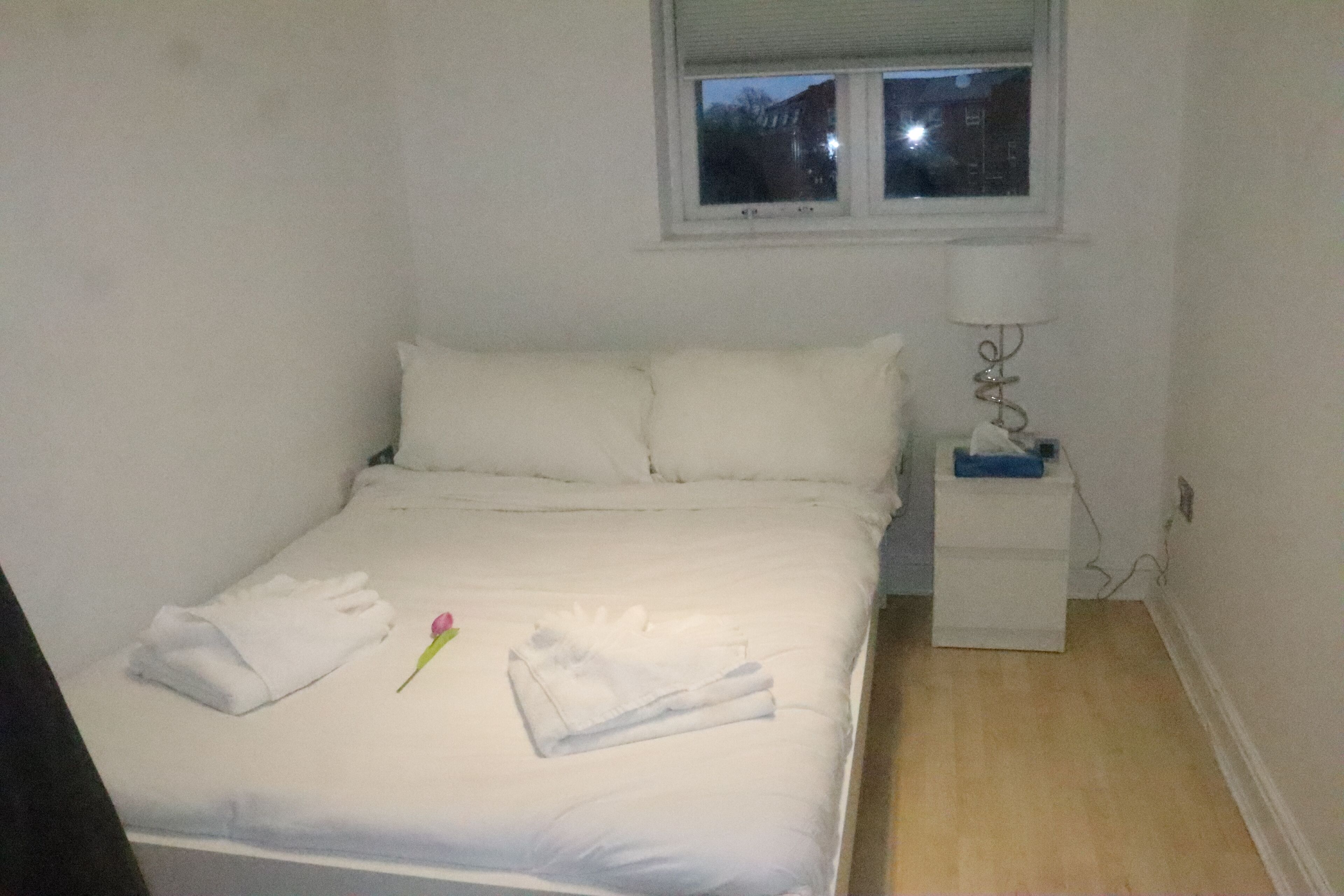 Una cama pequeña y ordenada con sábanas y almohadas blancas, una mesita de noche con lámpara, y una ventana sobre la cama.
