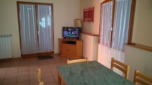 Quarto com uma mesa de jantar, cadeiras, uma televisão sobre um armário de madeira e uma janela com cortinas.