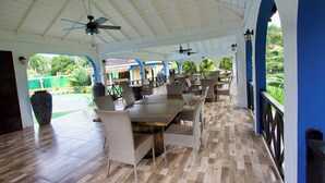 Une terrasse extérieure couverte, dotée d’une longue table et de chaises en bois, d’un ventilateur de plafond et offrant une vue sur un jardin.