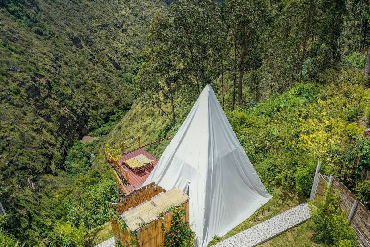 A white tent with a wooden deck and a small building with a red roof, surrounded by lush greenery and hills.