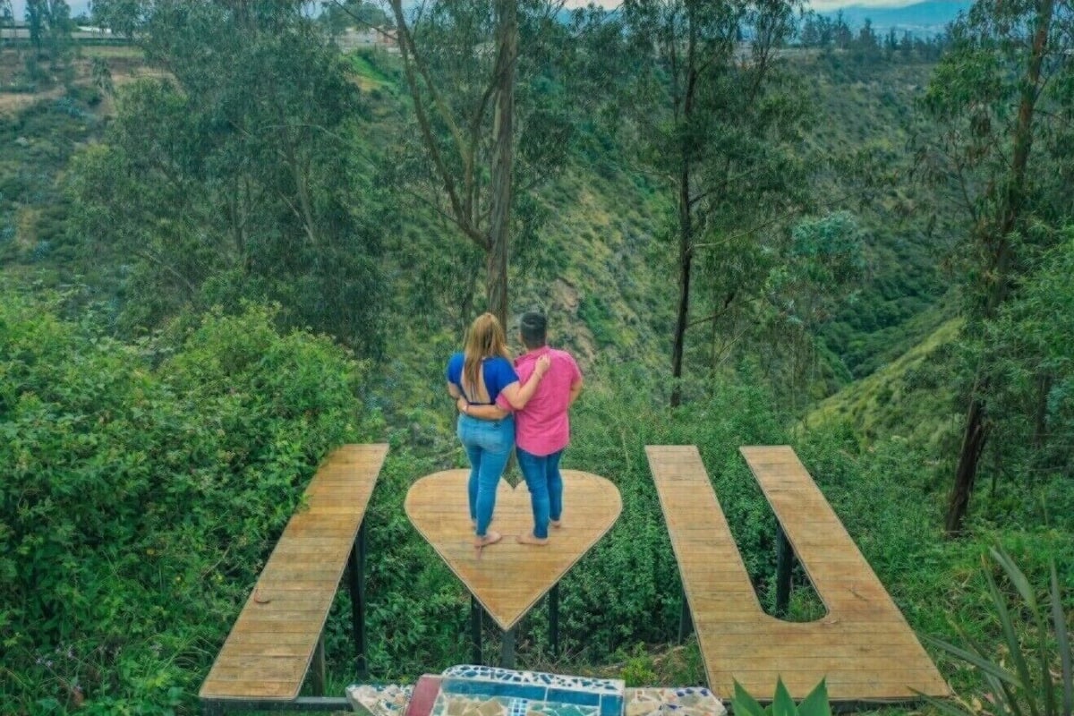 A couple standing on a heart-shaped wooden platform with a view of a lush green valley.