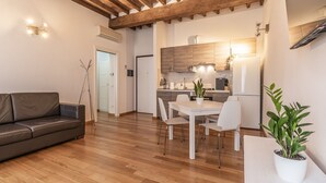 A modern living room with a wooden floor, a dark leather sofa, a dining table with white chairs, and a kitchen in the background. - [Piazza Martiri]-In pieno centro sotto al Castello (Carpi)