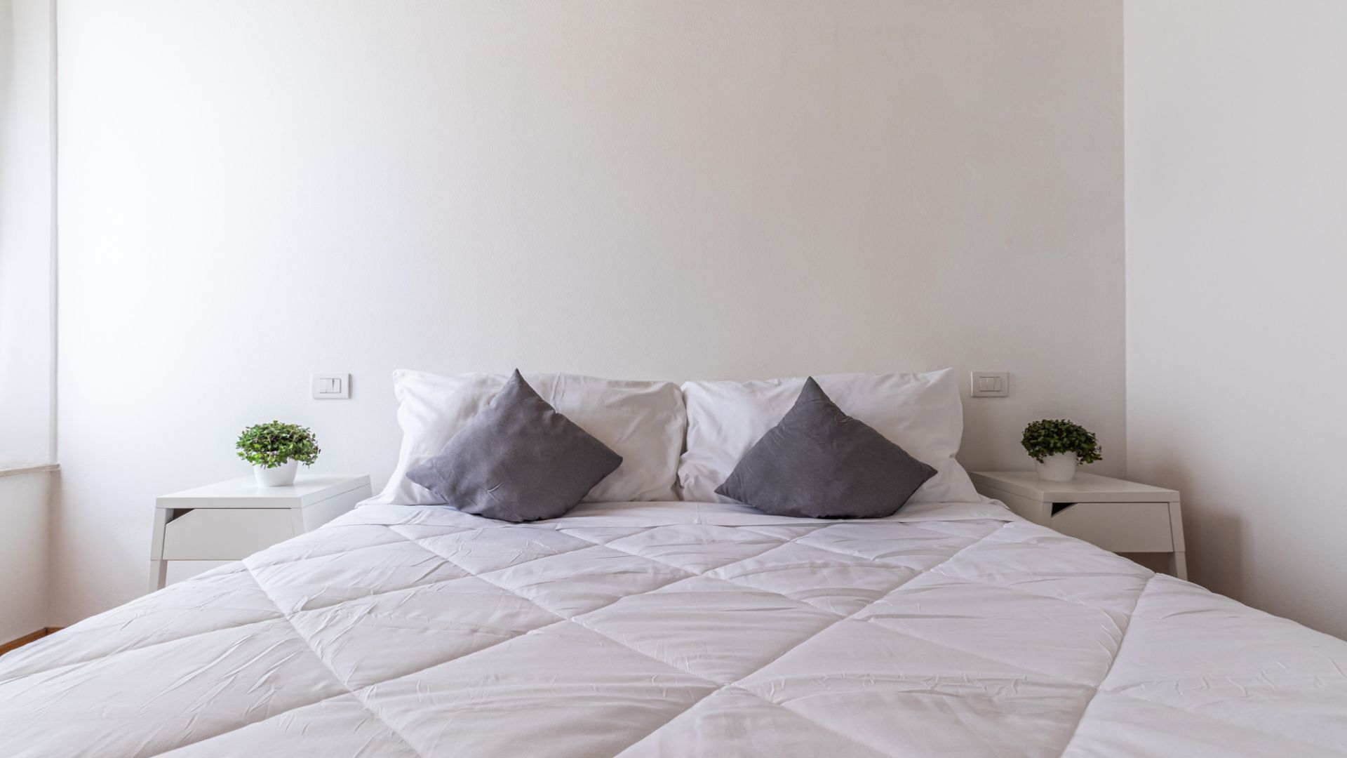 A bed with a white quilted cover, two gray pillows, and two bedside tables with small potted plants.