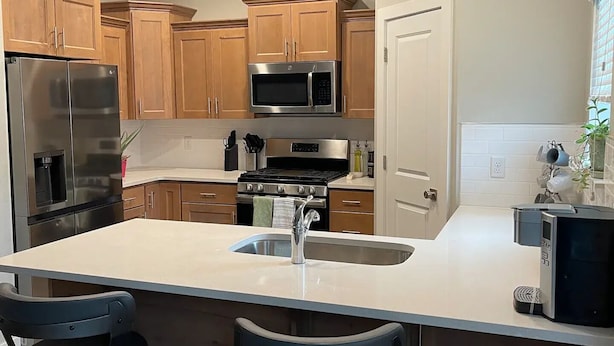 A modern kitchen with wooden cabinets, stainless steel appliances, a central island with a sink, and two bar stools. - Cute Modern townhouse close many Utah national parks (Cedar City)