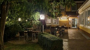 Un patio al aire libre con mesas y sillas, un seto y un edificio con la leyenda 'Motel' al fondo.