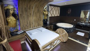 A modern bedroom with a wooden bed, a large mirror, a jacuzzi, and a mannequin. - Troia Motel (Foz do Iguaçu)