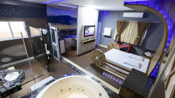 A modern bathroom with a large bathtub, a separate shower area, a vanity with a sink, and a television mounted on the wall. - Troia Motel (Foz do Iguaçu)