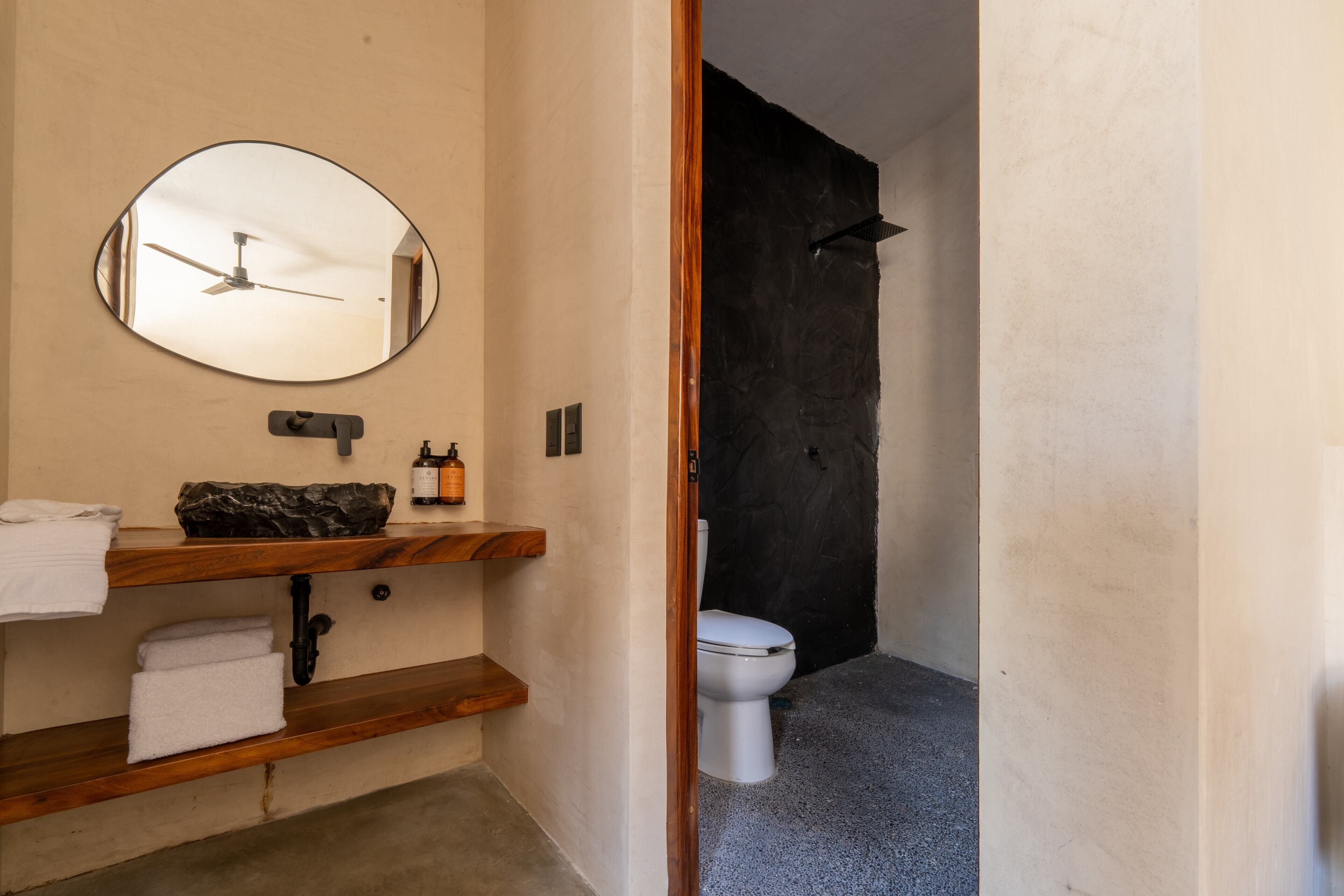 A bathroom with a wooden vanity, a round mirror, a toilet, and a shower area.