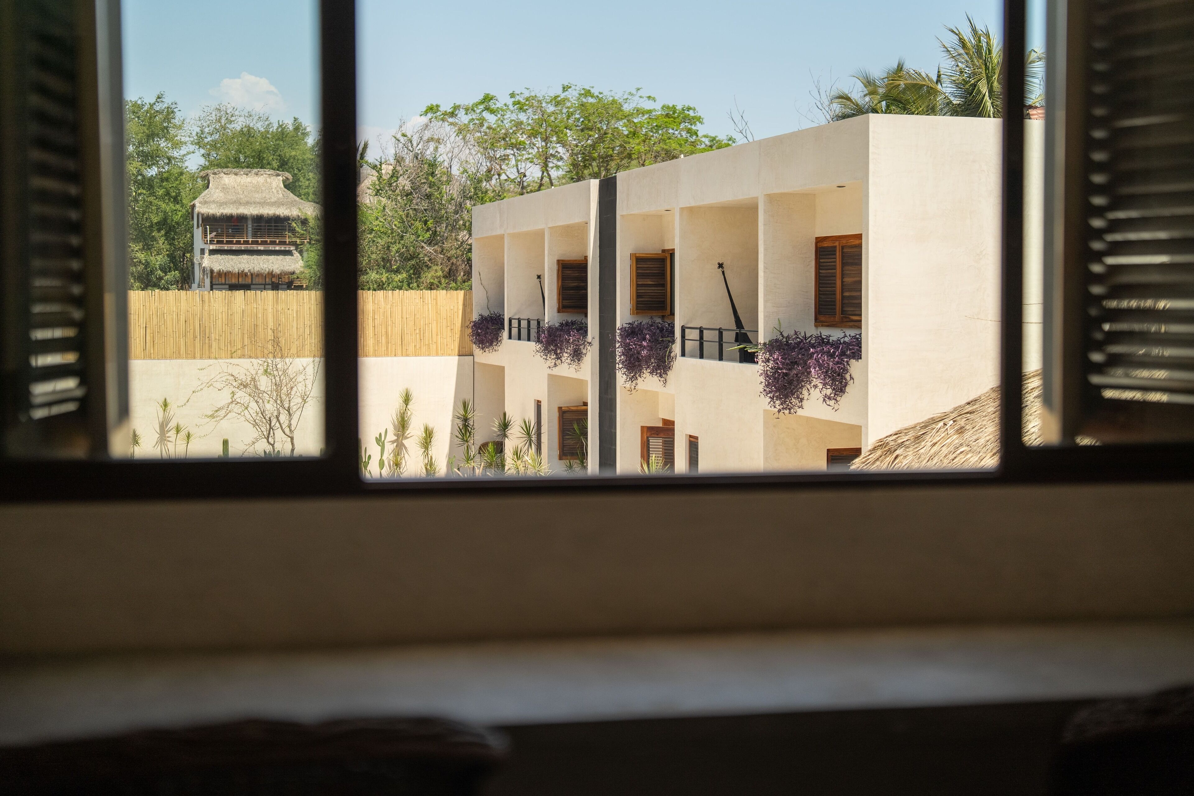 A view through a window of a building with multiple balconies, some with hanging plants, and a traditional thatched-roof structure in the background.
