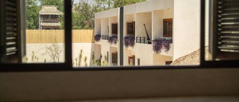 A view through a window of a building with multiple balconies, some with hanging plants, and a traditional thatched-roof structure in the background.
