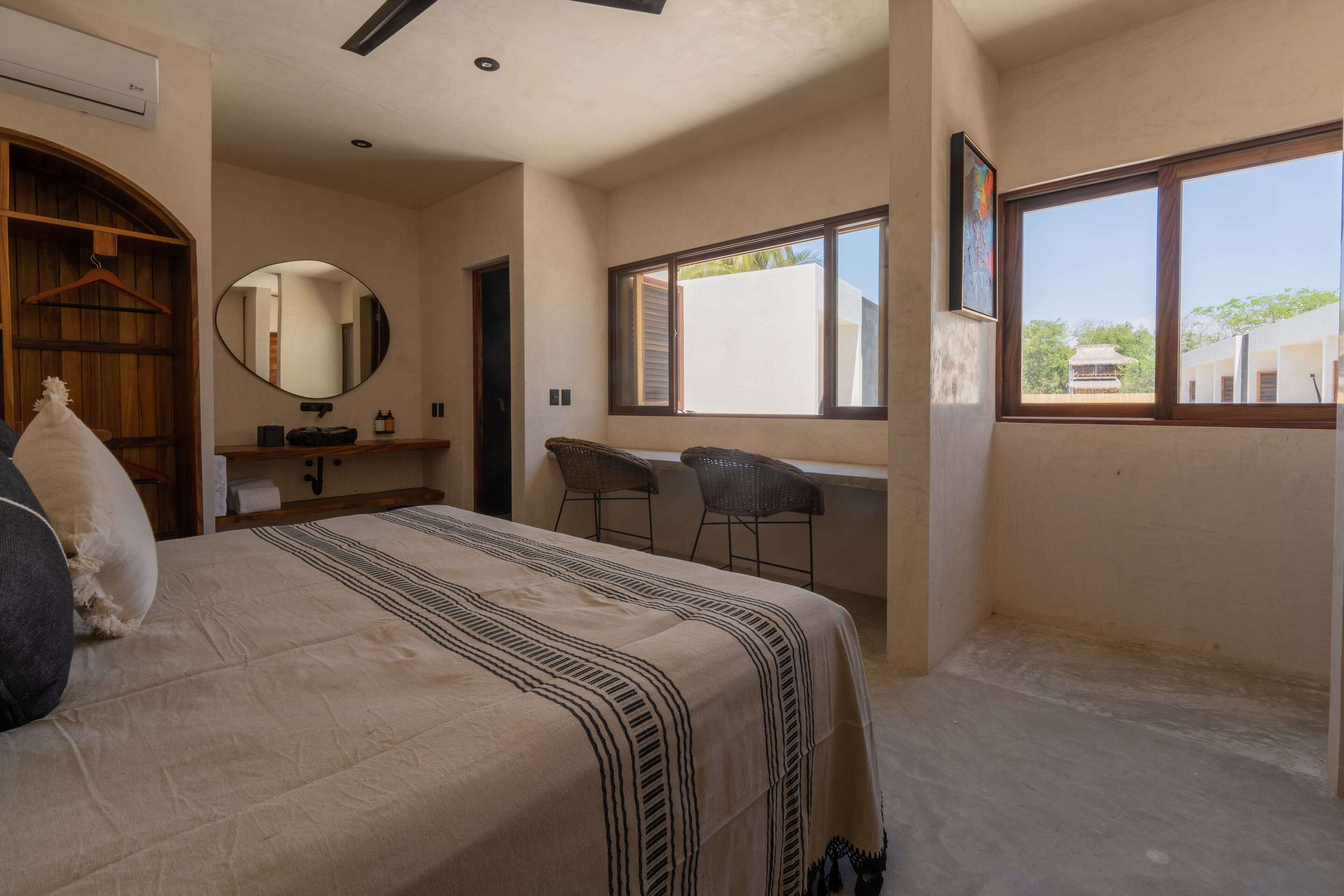 A bedroom with a bed, a desk, two chairs, a mirror, and a window.