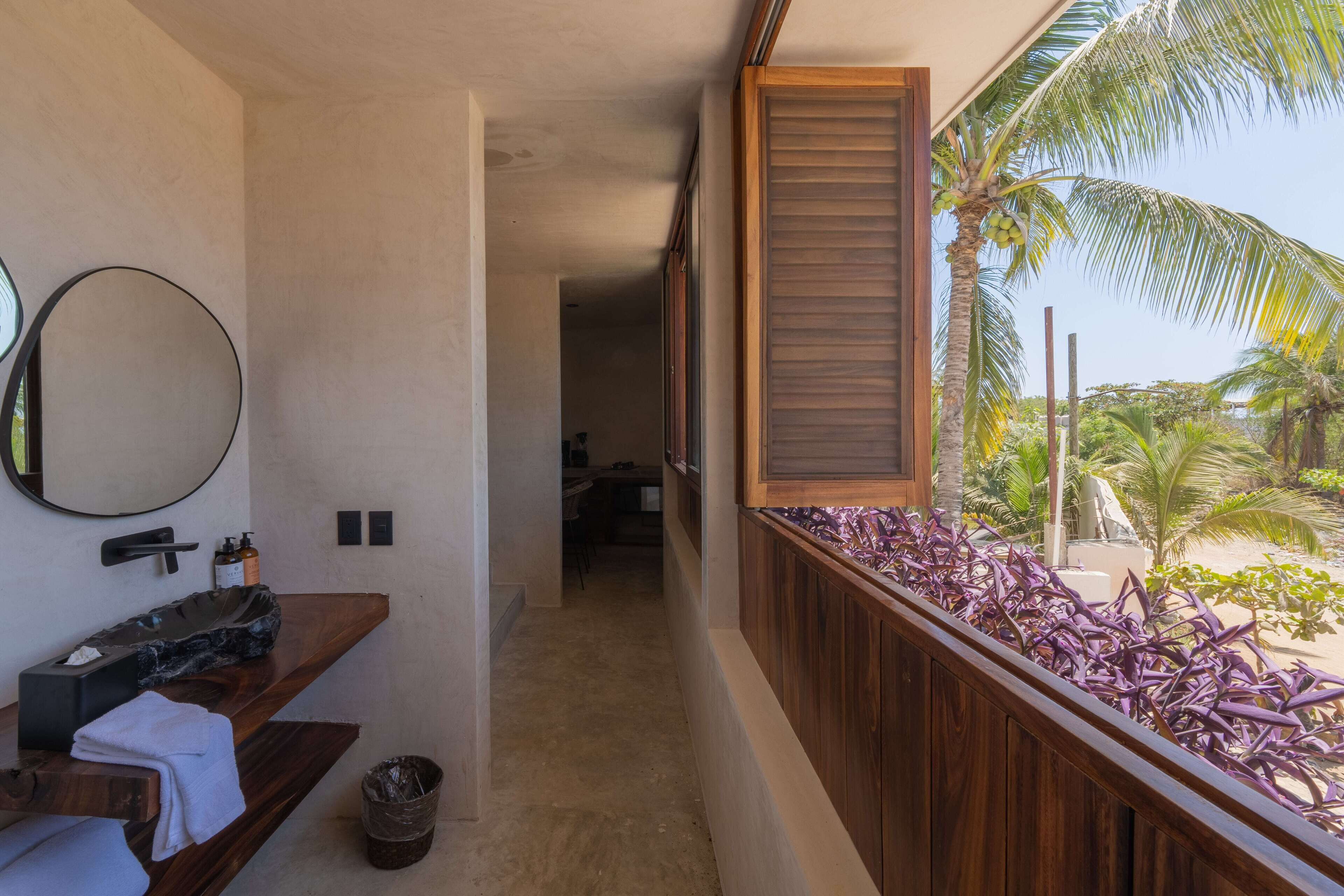 Un baño con tocador de madera, un espejo redondo y vistas a una playa con palmeras.