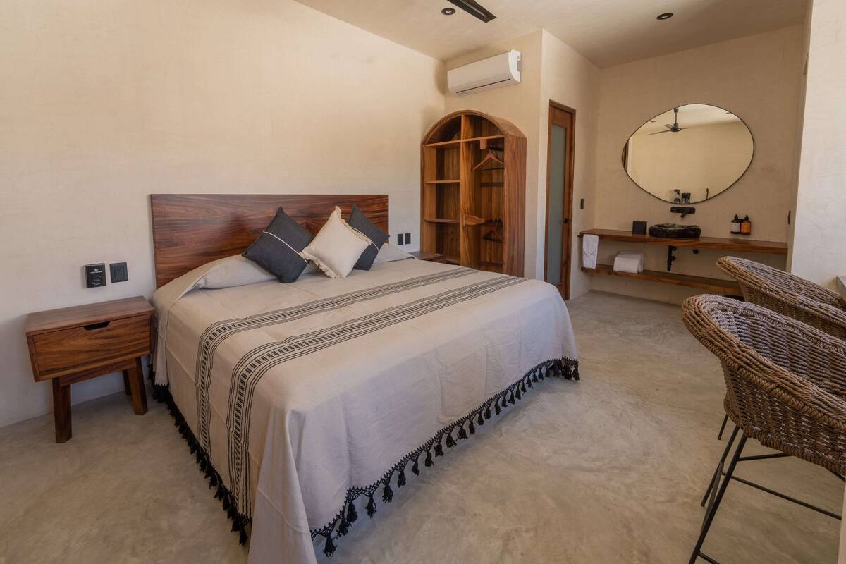 Une chambre avec une tête de lit en bois, un lit recouvert d’une couvre-lit à rayures noires et blanches, une armoire en bois, un miroir rond au-dessus du lavabo et des chaises en osier.