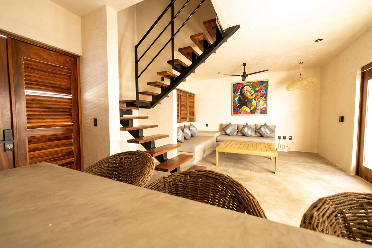 Un salon moderne avec un escalier en bois, un ensemble de canapés, des meubles en osier et un ventilateur de plafond.