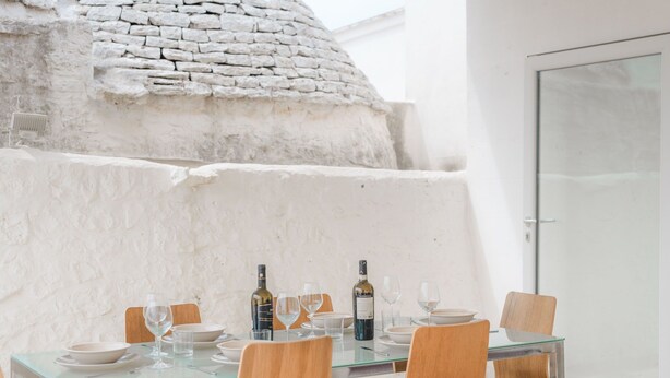 Une table à manger, avec des chaises, des assiettes, des verres et des bouteilles, devant un mur en pierre et une porte en verre à l’arrière-plan.