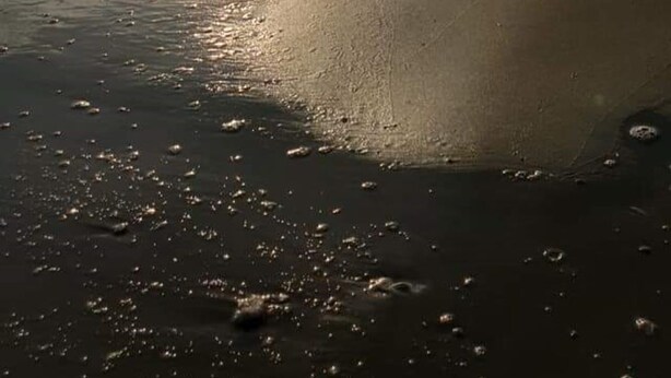 A beach with the sun low on the horizon, waves gently lapping at the shore, and buildings in the distance.