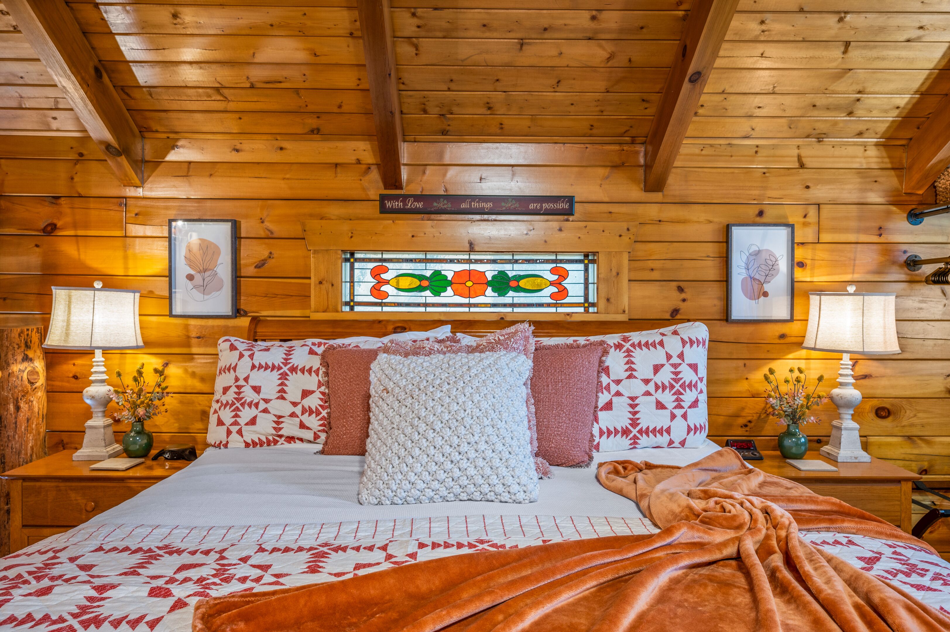 A wooden cabin bedroom with a bed, pillows, a blanket, a sign above the bed, and a stained glass window.