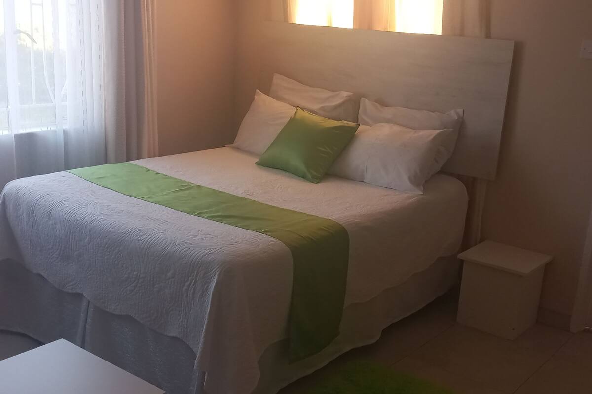 A neatly made bed with a white and green bedspread, a small bedside table, and a green rug.