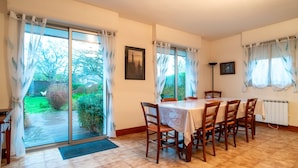 A dining area with a table and chairs, large windows with curtains, a framed picture on the wall, and a radiator.