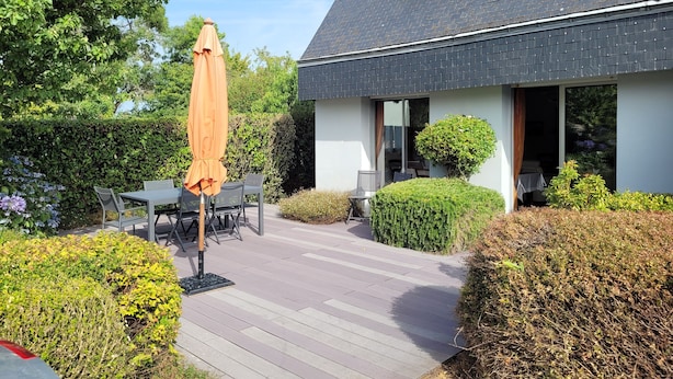 A modern house with a dark roof, a patio with a table and chairs, and a large umbrella.