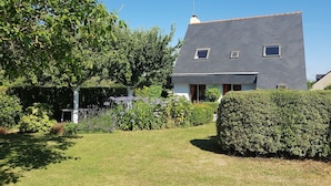 A modern two-story house with a sloped roof, a chimney, and a small garden with a hedge and flowering plants.