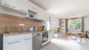 Eine moderne Küche mit weißen Schränken, einer Spülmaschine, einem Backofen und einem Waschbecken. Es gibt eine Essecke mit Tisch und Stühlen sowie ein Fenster mit Blick auf Grünflächen.