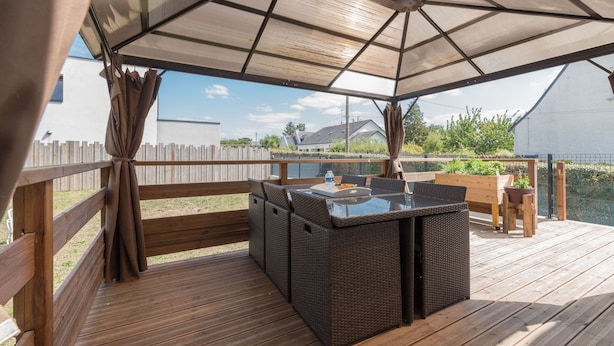 Eine Terrass mit überdachter Sitzecke, Holzdeck und Blick auf ein Haus und Grünanlagen im Hintergrund.