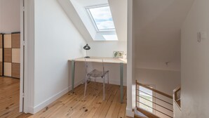 Ein Zimmer mit Schräge, einem Dachfenster, einem Holztisch, einem klaren Kunststoffstuhl und einer Treppe.