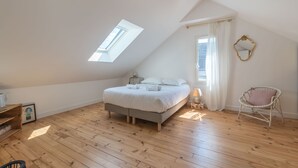 Ein Schlafzimmer mit schrägen Wänden, einem Oberlicht, einem Bett mit weißer Bettwäsche, einem Korbstuhl und einem Spiegel an der Wand.