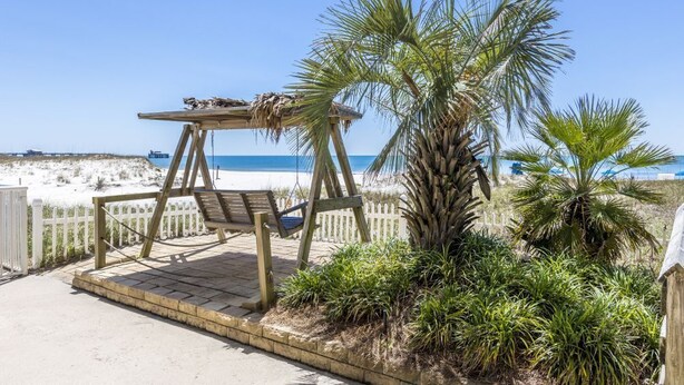 Une balançoire en bois avec un banc, entourée de palmiers et d’une clôture blanche, donnant sur une plage.