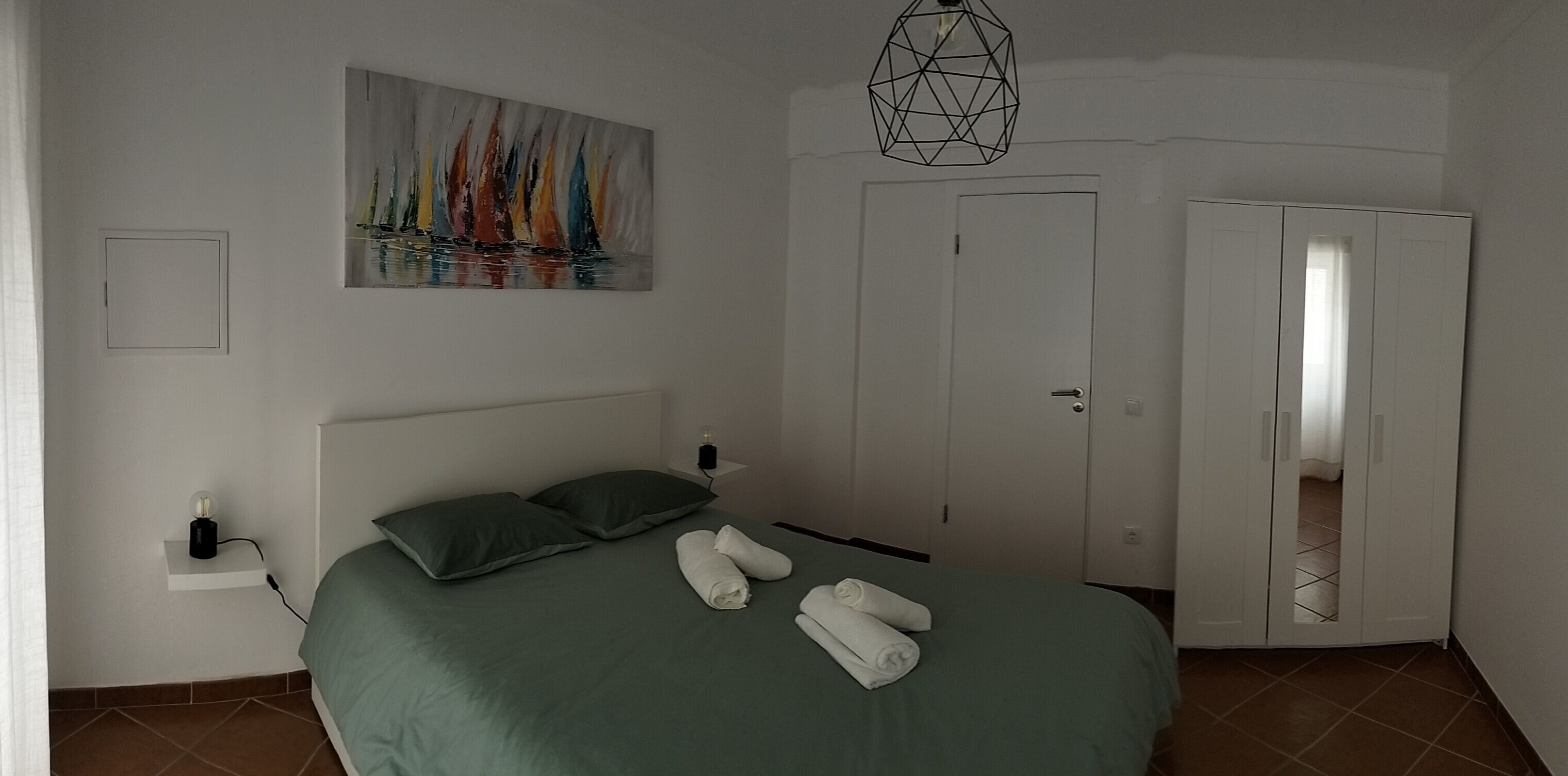 A neatly made bed with pillows and towels, a painting of sailboats on the wall, a geometric light fixture, and a white wardrobe.