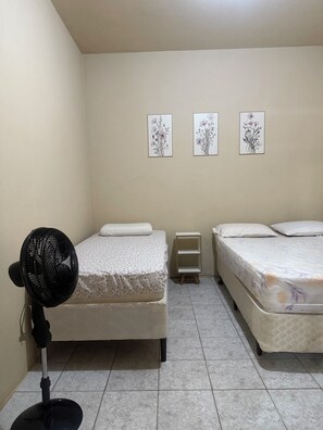 A bedroom with two beds, a standing fan, and three framed floral artworks on the wall. - Sítio Aconchegante em Amparo/sp com uma Linda Vista Para Apreciar o pôr do sol (AMPARO)