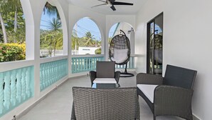 A balcony with wicker furniture, a ceiling fan, and a view of palm trees and a white fence. - Spacious Home with pool, steps from the beach in Puerto Rico! (Vega Alta)