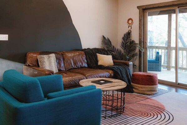 Ein Wohnzimmer mit braunem Ledersofa, blauen Sesseln, einem runden Couchtisch und einem großen Fenster mit Blick ins Freie.