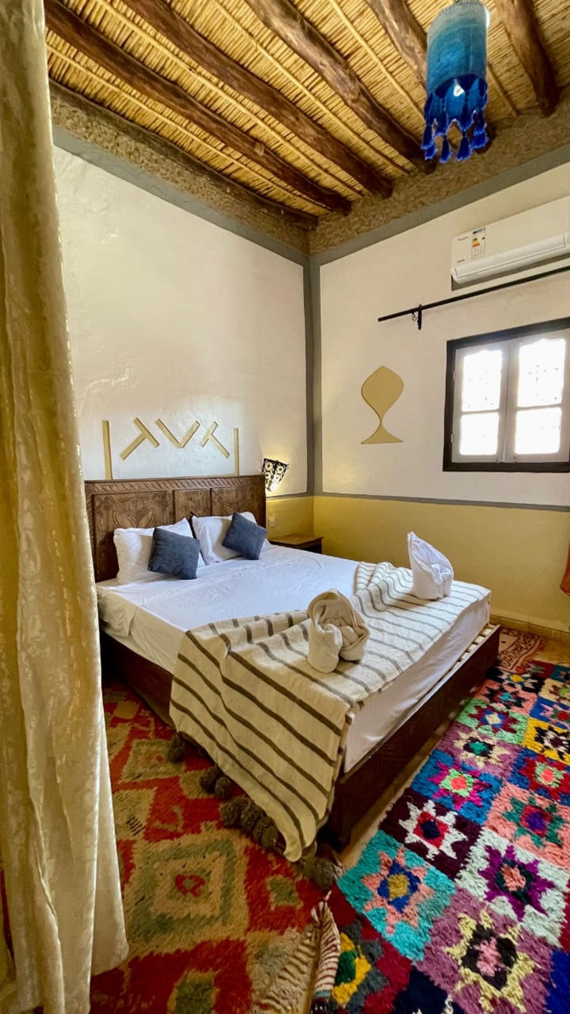Une chambre à coucher avec un lit, des oreillers, une couverture, un tapis coloré, un plafond en bois et un appareil de climatisation.