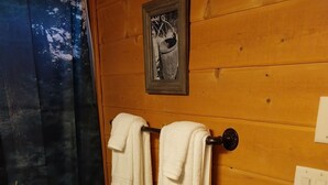 A wooden-paneled bathroom with two white towels hanging on a towel rack, a framed picture above the rack, and a red bath mat on the floor. - Miners Cabin - New Private and Cozy (Cottonwood)