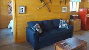 A cozy cabin interior with a blue sofa, wooden walls, a red refrigerator, and a small kitchen area. - Miners Cabin - New Private and Cozy (Cottonwood)