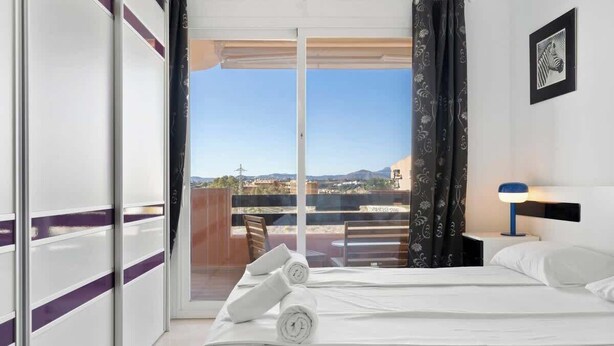 A hotel room with a bed, a nightstand, a lamp, a framed picture, and a view of the outdoors. - Torreblanca with pool by Costarentals - Ref 63 (Fuengirola)