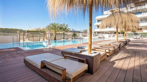 A poolside area with lounge chairs, a thatched umbrella, and a view of a building in the background. - La Cala de Mijas Modern Flat with Pool - ref 240 (Las Lagunas de Mijas)