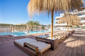 Zona de piscina con sillones de descanso, una sombrilla de paja y vista a un edificio al fondo.