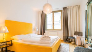 Ein Hotelzimmer mit einem gelben Bett, einem Nachttisch mit Lampe, einem Sessel, einem Fenster mit Vorhängen und einem Spiegel.