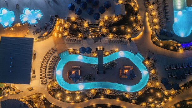 Vista aérea de un complejo turístico con una piscina serpenteante, áreas de descanso y mobiliario exterior.