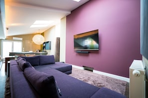 A modern living room with a large sectional sofa, a flat-screen TV mounted on the wall, and a kitchen in the background.