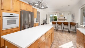 A modern kitchen with wooden cabinets, stainless steel appliances, and a large island with bar stools. - The Desert Flower, designer home in gated community/ private pool and jacuzzi. (Palm Springs)