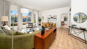 A spacious living area with a sofa, coffee table, and a wooden console table. There is a dining area with chairs and a table, and a large window with curtains. - The Desert Flower, designer home in gated community/ private pool and jacuzzi. (Palm Springs)