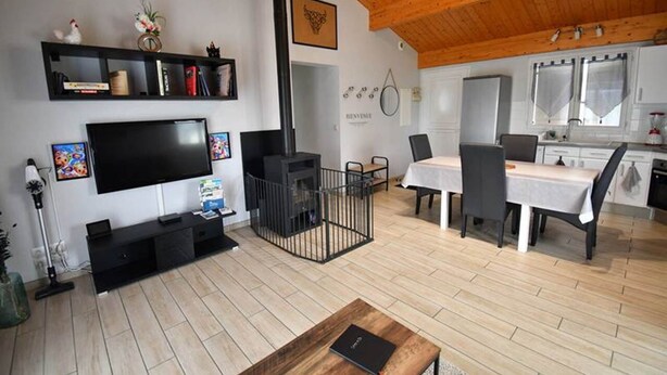 A modern living room with a flat-screen TV, a fireplace, a dining area with a table and chairs, and a kitchen in the background.
