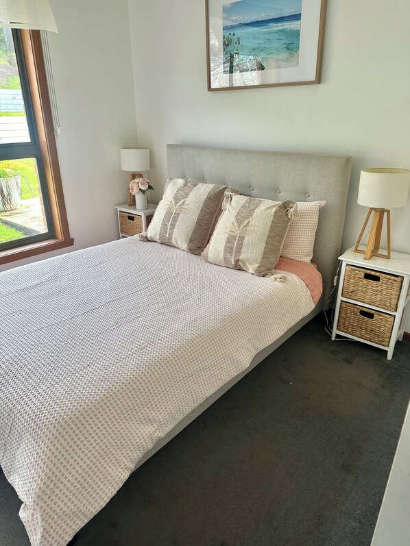A neatly made bed with a patterned bedspread, two bedside tables with lamps, a framed picture on the wall, and a window with a view of greenery. - Rye Beach Escape (Rye)