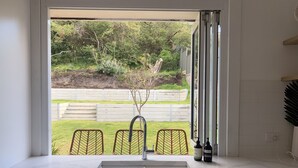 A kitchen with a white sink, a window overlooking a garden, and a view of a wooden fence. - Rye Beach Escape (Rye)