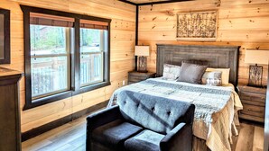 A wooden cabin bedroom with a bed, a chair, a ceiling fan, and a window with a view of the outdoors.